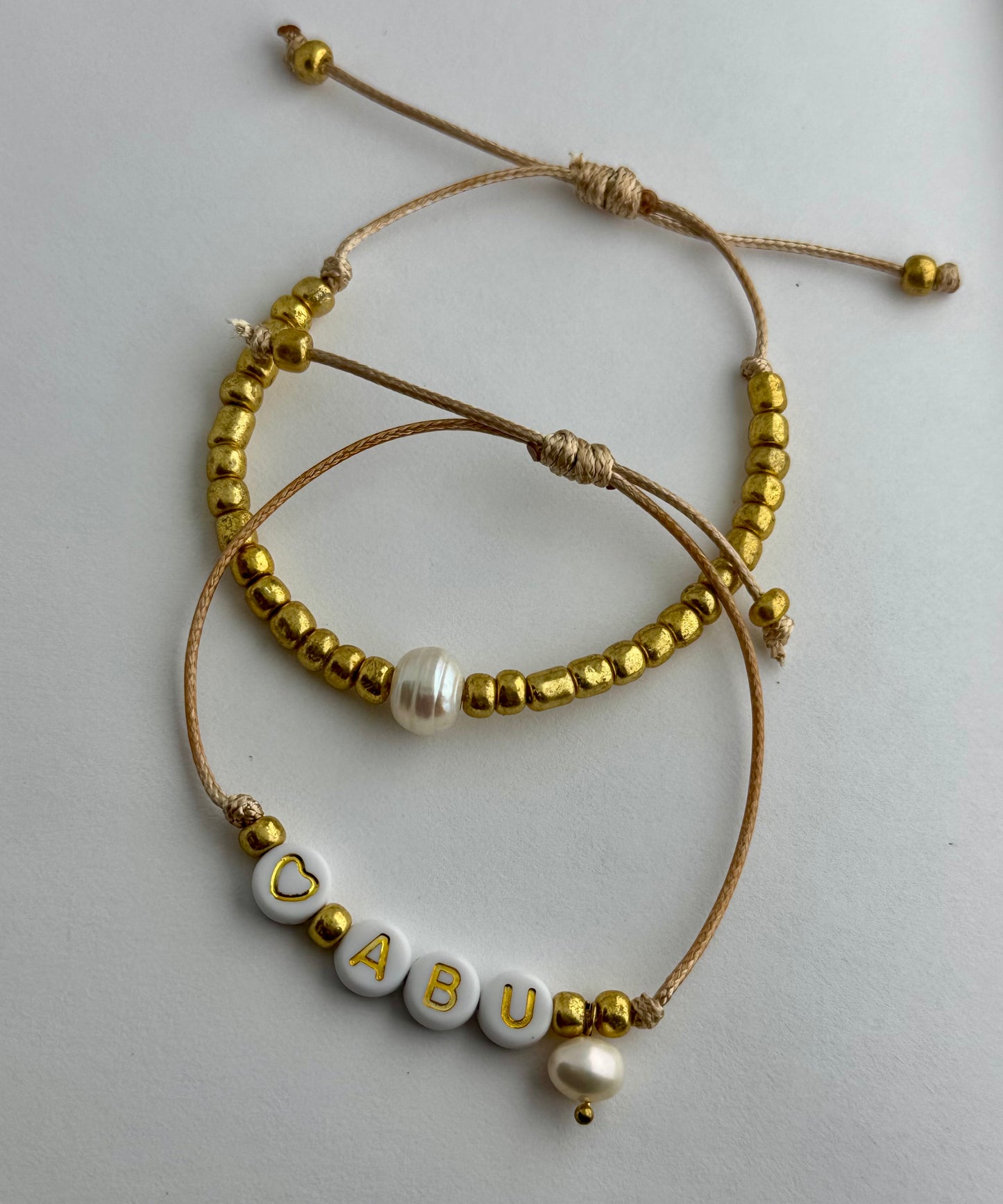 ABU - Dúo de pulseras en hilo hecha a mano color dorado con la palabra ABU, perlas de agua dulce y mostacillas.