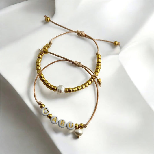 ABU - Dúo de pulseras en hilo hecha a mano color dorado con la palabra ABU, perlas de agua dulce y mostacillas.